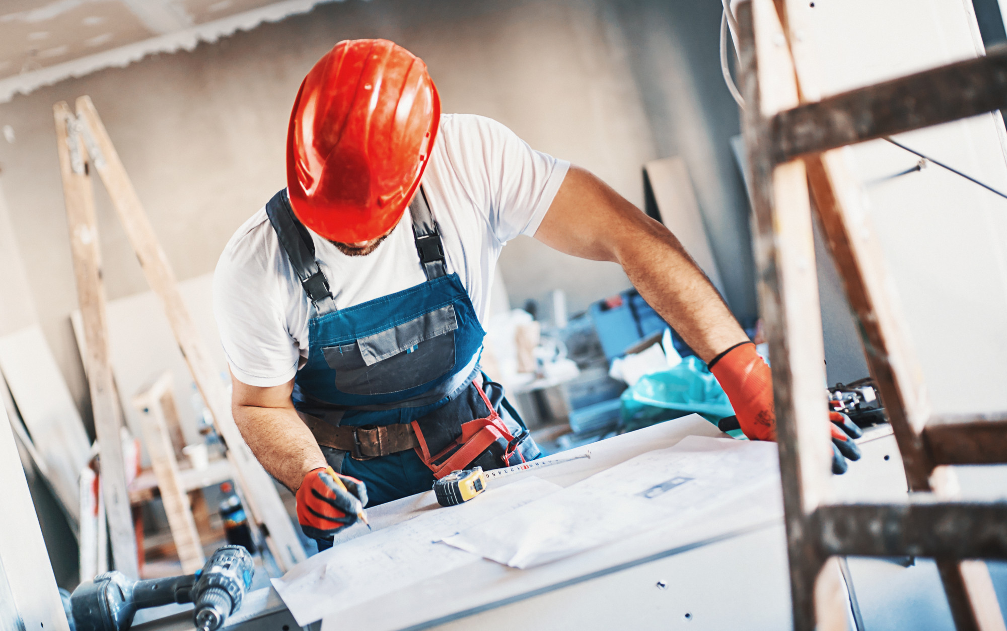 Construction worker going through blueprints at a construction site. there are some uncertanties he;s trying to solve. He's wearing red work helmet and gloves and blue uniform. There are tools next to him.